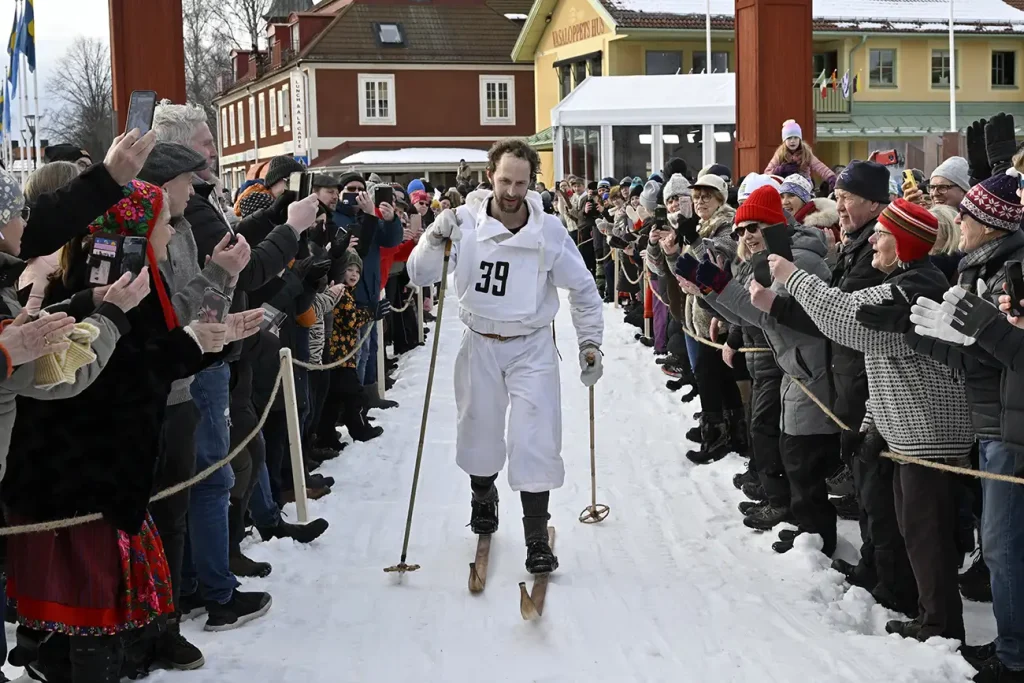 Skidåkaren Anders Södergren i vit gammaldags skiddräck går i mål på en andraplats i Jubileumsvasan 2022 samtidigt som han påhejas av en stor publik runt spåret in mot mål.