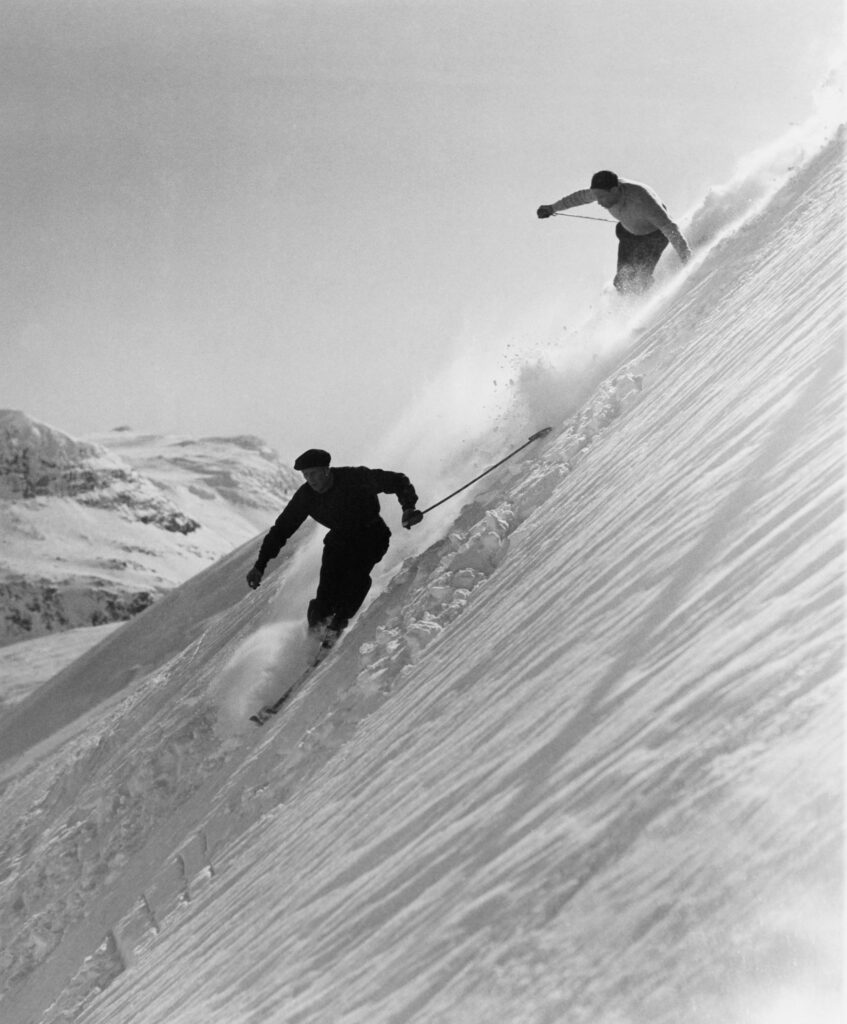 A 60-year-old Olle Rimfors makes a ruade turn on a wind packed snow with his 205 cm wooden skis on the Nordalsbranten off-piste run in 1956. Following on his track is a student who just got his ski instructor diploma. Photo: Sven Hörnell