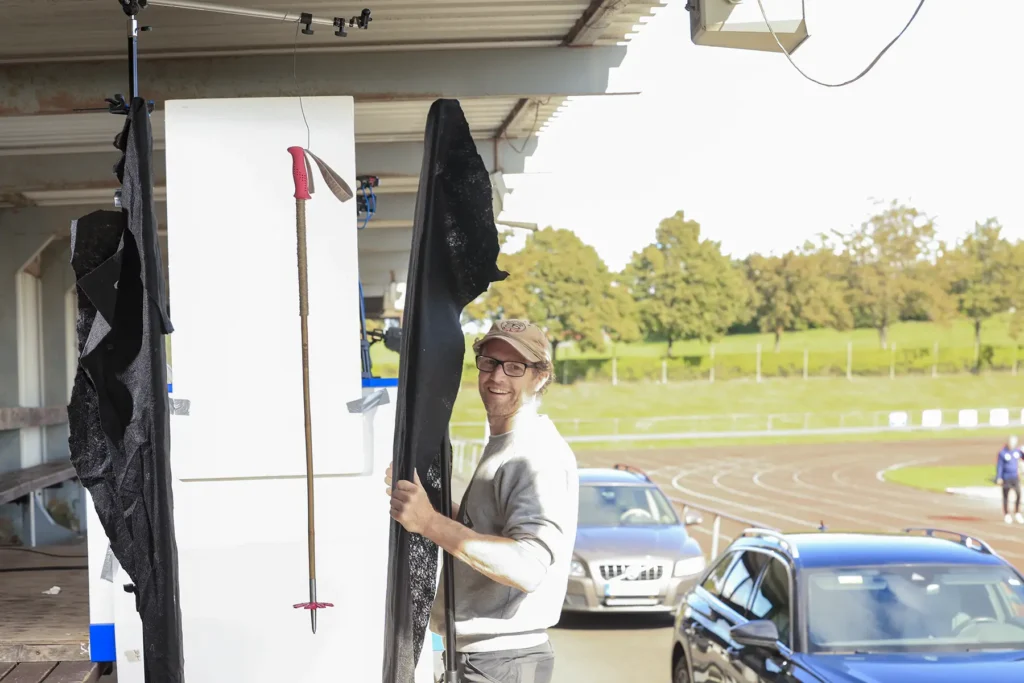 Fabian Rimfors holds up a black screen next to a bamboo ski pole being photographed in a sports grandstand.