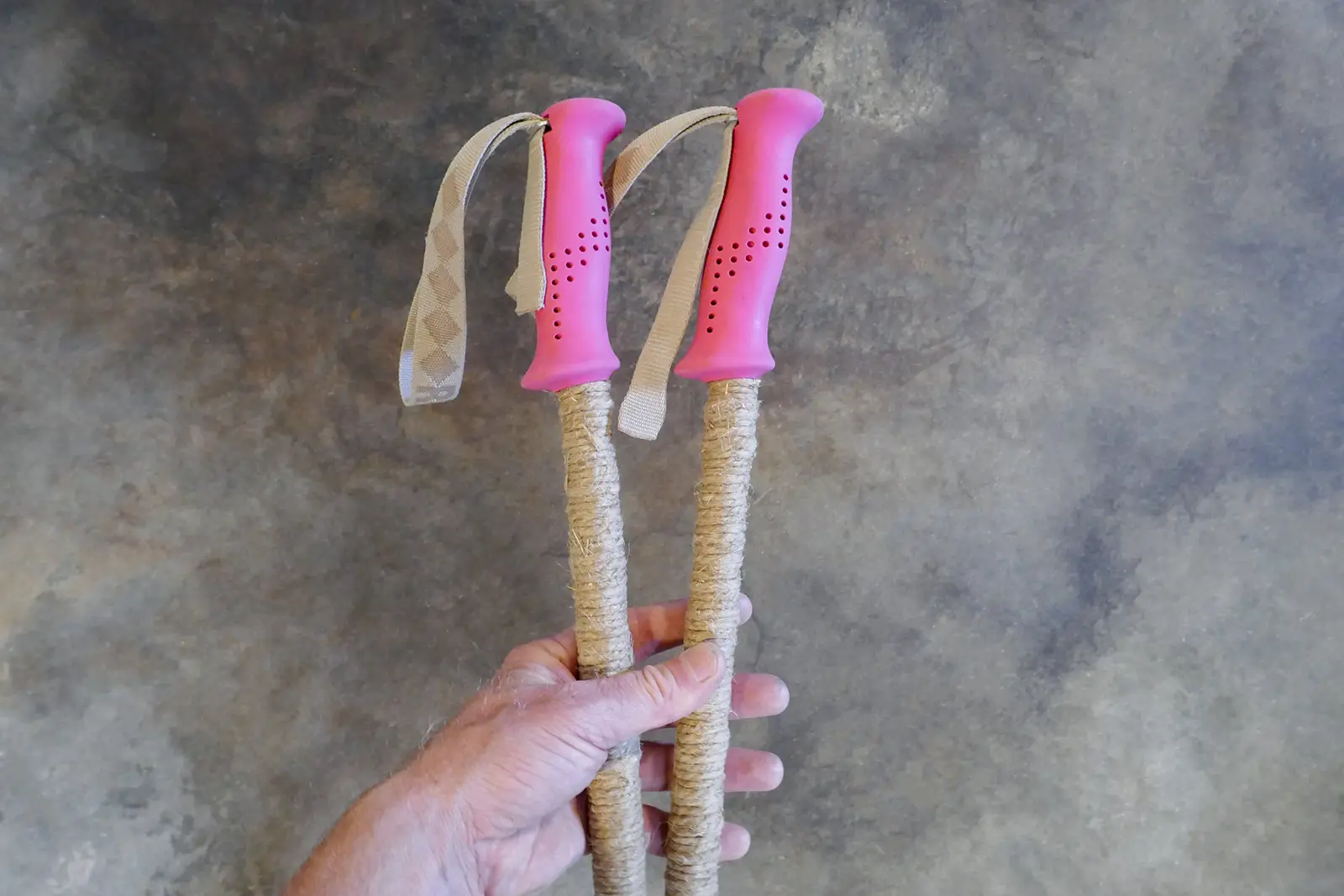 Hand holding a pair of ski poles with pink grips and long grips of wrapped hemp cord.