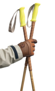 Hand with brown leather glove holds two bamboo ski poles with fluo yellow grips and reindeer leather grip extensions.