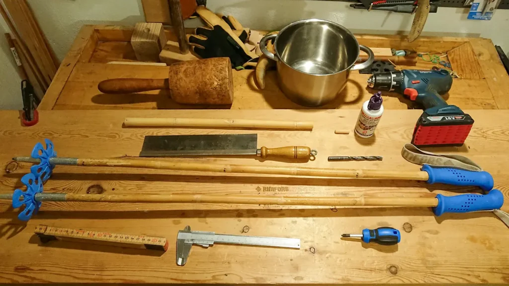 Blue bamboo ski poles with the tools needed to lengthen the poles; carpenter's ruler, caliper, wood glue, drill bit, drill, fine tooth saw, screwdriver, pot and a piece of bamboo.