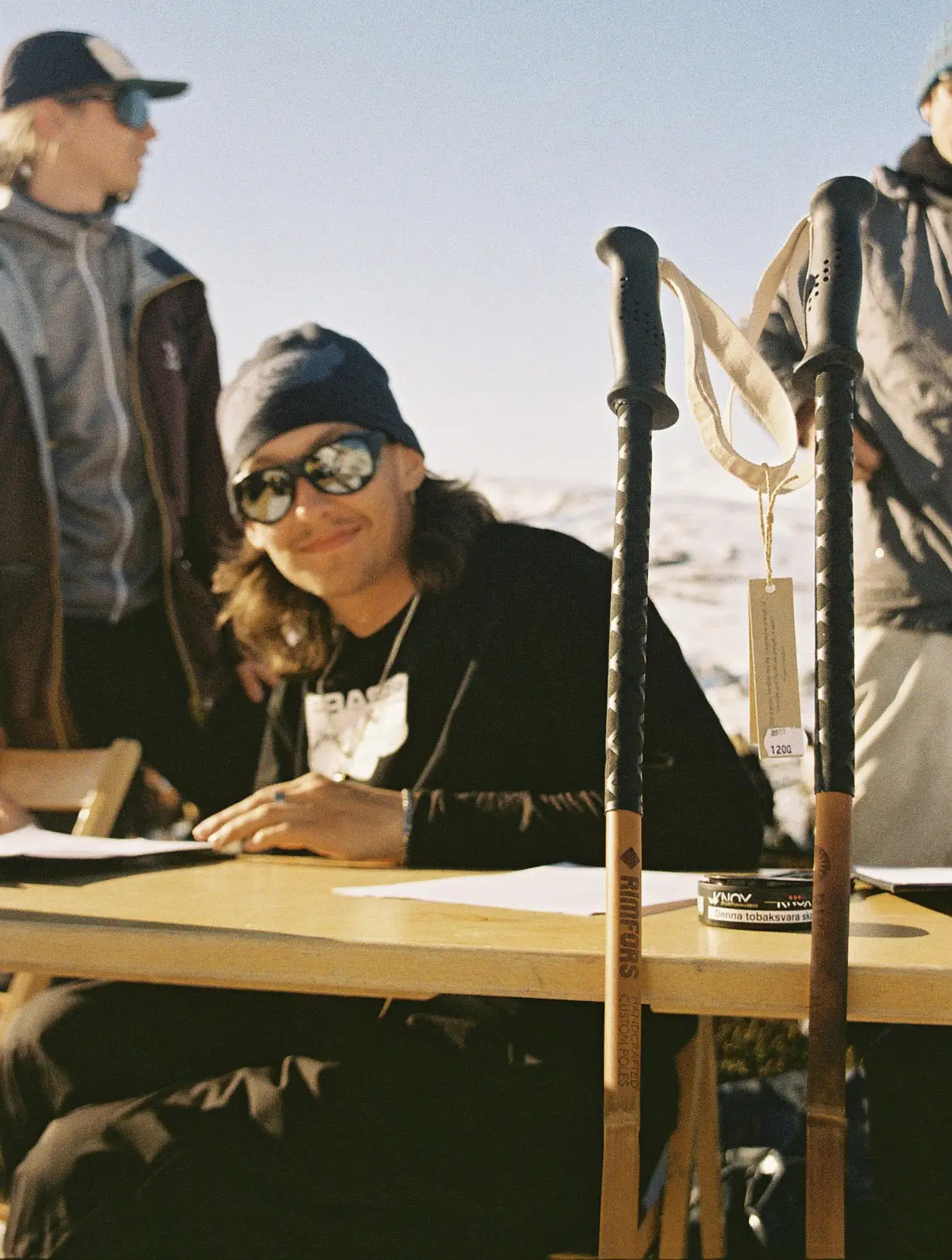 A guy with long brown hair, a beanie, and sunglasses sitting at a table with a pair of bamboo ski poles.