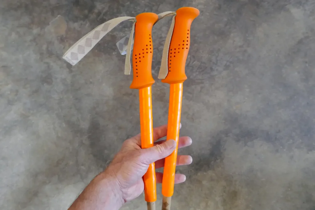 Hand holding a pair of ski poles with orange grips and grip extensions of orange overgrip tape.