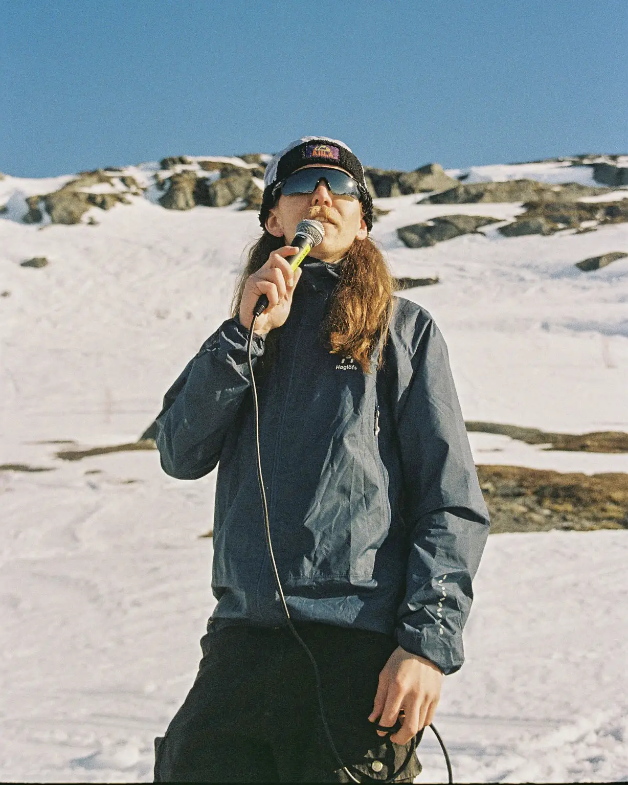 A long-haired dark blonde guy with sunglasses and a blue jacket speaks into a microphone in front of a snow-covered mountain.