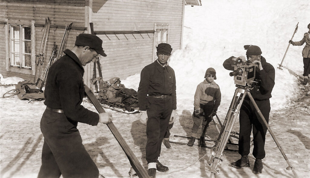Olle Rimfors vallar skidor medan fotografen Per Flood ställer in kameran under inspelningen av Sveriges första skidfilm – Tre män på skidor. Gunnar Dyhlén står i mitten och regisserar.