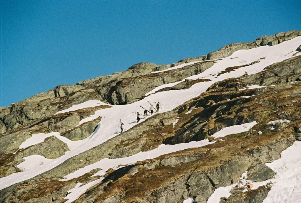 Skiers carrying their skis on their shoulders as they ascend a steep, narrow snowfield surrounded by bare cliffs in the sun.