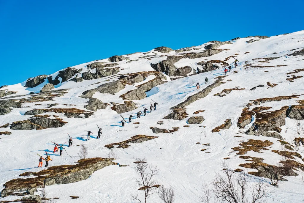 18 skidåkare och snowboardåkare på väg upp för ett berg till fots.