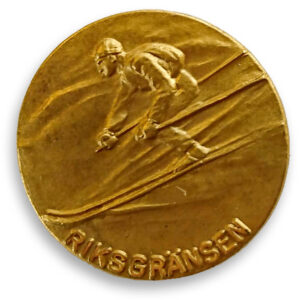 The Riksgränsen Downhill badge in gold, with a skier going downhill a mountain.